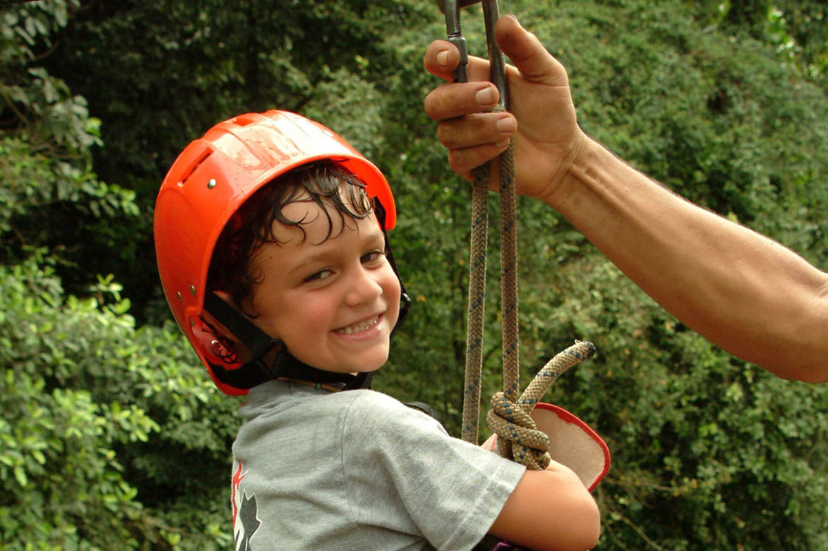 The hand of a guide holds the zip line while a little boy smiles at the camera